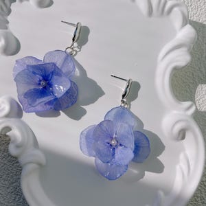 Peut inclure: Une paire de boucles d'oreilles en forme de fleurs d'hortensia bleu clair. Chaque boucle d'oreille présente un groupe de pétales avec une petite perle centrale. Les boucles d'oreilles sont suspendues à des tiges argentées et sont présentées sur un plateau blanc décoratif.