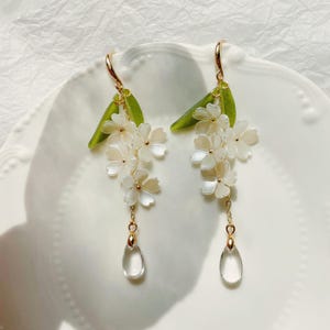 May include: Dangle earrings with white flower charms, gold accents, and clear teardrop pendants. The earrings have green leaf details and gold-coloured ear wires. The earrings are displayed on a white plate. The earrings are a delicate floral design.