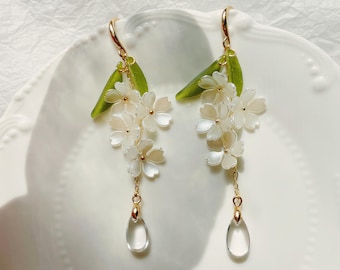 Könnte beinhalten: Ohrringe mit weißen Blüten, goldenen Akzenten und transparenten Tropfenanhängern. Die Ohrringe haben grüne Blattdetails und goldfarbene Ohrhaken. Die Ohrringe sind auf einem weißen Teller präsentiert. Ein filigranes florales Design.