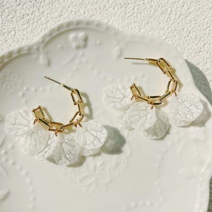 May include: A pair of gold-toned hoop earrings with a chain design. Each earring features three white, petal-shaped lace accents, creating a delicate and feminine aesthetic. The earrings are displayed on a white, decorative plate.