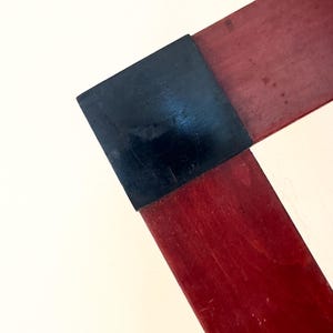 Op de afbeelding: Close-up van een hoek van een houten frame. Het frame is gemaakt van donkerrood hout en heeft een zwart vierkant op de hoekverbinding. De achtergrond is effen gebroken wit.