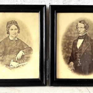 Könnte beinhalten: Zwei schwarze gerahmte Porträts eines Mannes und einer Frau in antiken Rahmen. Die Frau trägt ein dunkles Kleid und hat ihr Haar nach hinten gebunden. Der Mann trägt einen dunklen Anzug und eine Fliege.