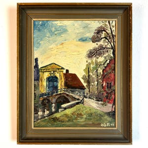 May include: Oil painting in a gold frame showing a European village. A yellow building with a blue arched doorway and a red-roofed structure are connected by a bridge. The sky is light blue and yellow, with trees and water in the foreground.