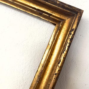 Op de afbeelding: Close-up van een hoek van een goudkleurige fotolijst. De lijst heeft een verweerde, antieke uitstraling met zichtbare slijtage. De gouden afwerking is licht vervaagd, waardoor hints van het onderliggende materiaal zichtbaar worden. De lijst staat tegen een witte, getextureerde muur.