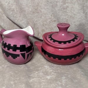 May include: A pink ceramic creamer and sugar bowl set. The creamer has a white interior and a handle. Both pieces are decorated with black geometric patterns. The sugar bowl has a lid with a small handle.