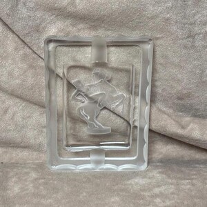 May include: A clear glass ashtray with a raised, etched design of a centaur. The ashtray has a square shape with rounded corners and a decorative border.