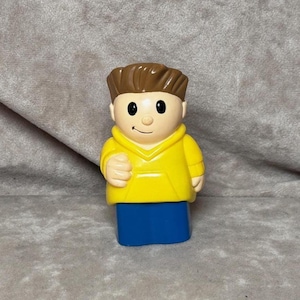 May include: A plastic toy figure of a person wearing a yellow hooded sweatshirt and blue pants. The figure has brown hair, black eyes, and a smiling face. The toy is designed in a cartoon style.