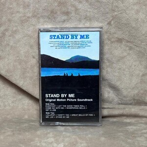 May include: A cassette tape with a blue and white image of a lake and trees. The text "STAND BY ME" is printed on the cassette tape. The text "Original Motion Picture Soundtrack" is printed below the title. The song titles "Everyday", "Let The Good Times Roll", "Come Go With Me", "Whispering Bells", "Get A Job", "Lollipop", "Yakety Yak", "Great Balls Of Fire", "Mr. Lee", and "Stand By Me" are listed on the cassette tape.