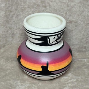 May include: A small, hand-painted ceramic vase with a white background and a purple, orange, and yellow sunset design. The vase has black geometric patterns around the top and bottom.