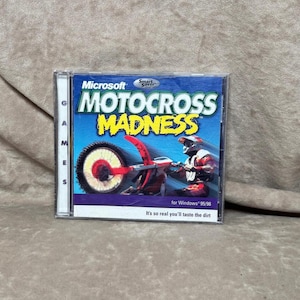 May include: A Microsoft Motocross Madness PC game in a clear plastic case. The cover features a motocross bike with a rider and the game's title in yellow and blue. The text "It's so real you'll taste the dirt" is at the bottom.