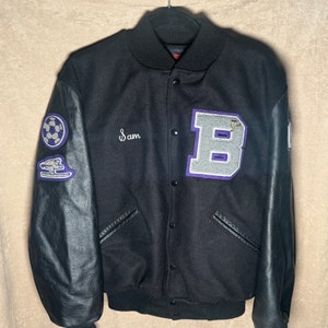 May include: Black varsity jacket with black leather sleeves. Features a large gray and purple letter "B" patch, a soccer ball patch, and a patch with a spaceship design. The name "Sam" is embroidered in cursive.