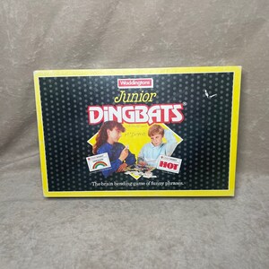 May include: A yellow, black, and white game box for the game "Junior Dingbats". The box features a black background with white stars and the game title in yellow and black. The game is described as "The brain bending game of funny phrases".
