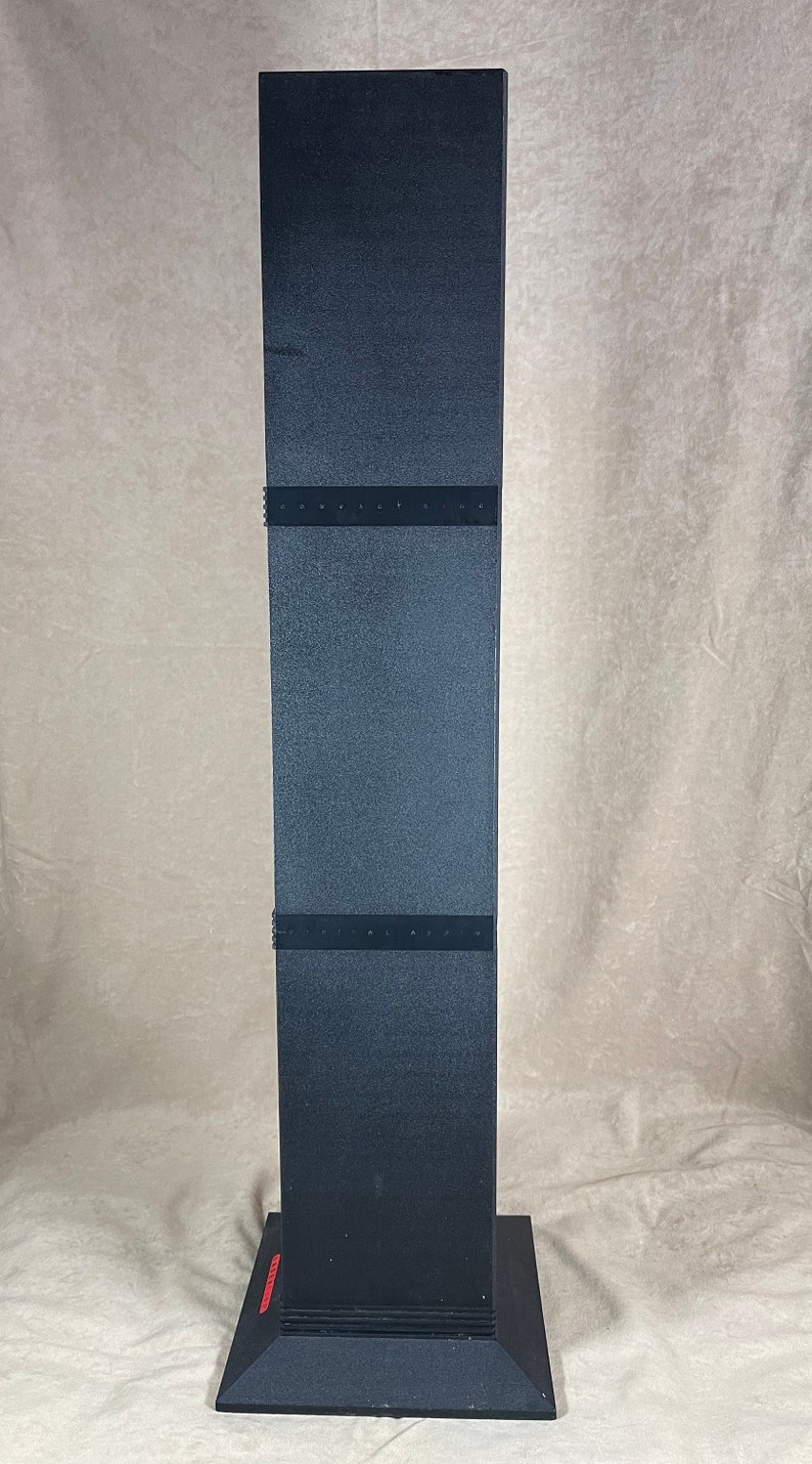 May include: A tall, black, rectangular display stand with a square base. The stand has a textured surface and is divided into three sections with dark bands. The base has a red accent.