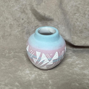 May include: A small, round, light blue and pink ceramic vase with a white geometric design.