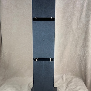 May include: A tall, black, free-standing display stand with a rectangular base and a vertical column. The column has two rectangular openings. The stand is made of a matte black material.