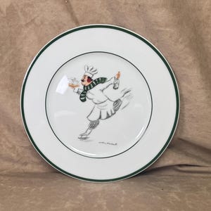 May include: White ceramic plate with a green rim and a whimsical illustration of a chef ice skating while holding a tray of food and a bottle. The chef is wearing a white uniform, a green and white scarf, and a chef's hat.