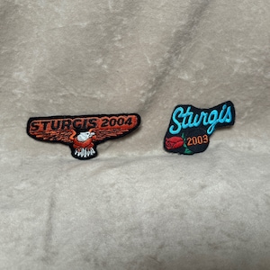 May include: Two embroidered patches, one with a red eagle and the text "Sturgis 2004" and the other with a blue cursive "Sturgis" and a red rose with the text "2003".