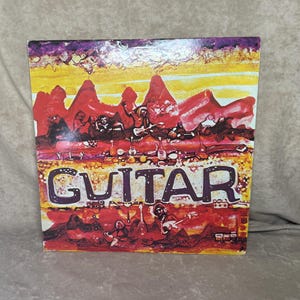 May include: Vintage vinyl record album cover with the word "GUITAR" in large, stylized letters. The artwork features a vibrant, abstract design with red, yellow, and purple hues, depicting musicians and instruments. The album cover is slightly worn.