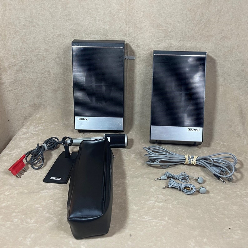 May include: Two black Sony speakers with silver accents, a black microphone with a stand, a black carrying case, and two sets of grey ear buds with cords.