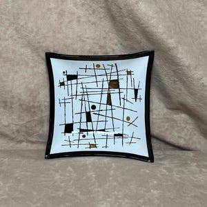 May include: Square decorative dish with a black rim and a light blue interior. The dish features a mid-century modern design with black lines, squares, and dots, along with brown and gold accents. The dish is a unique piece of home decor.