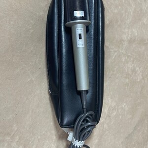 May include: A silver Sony F-915 microphone with a black mesh grille and a black leather carrying case. The microphone has a switch for on/off and a volume control knob.