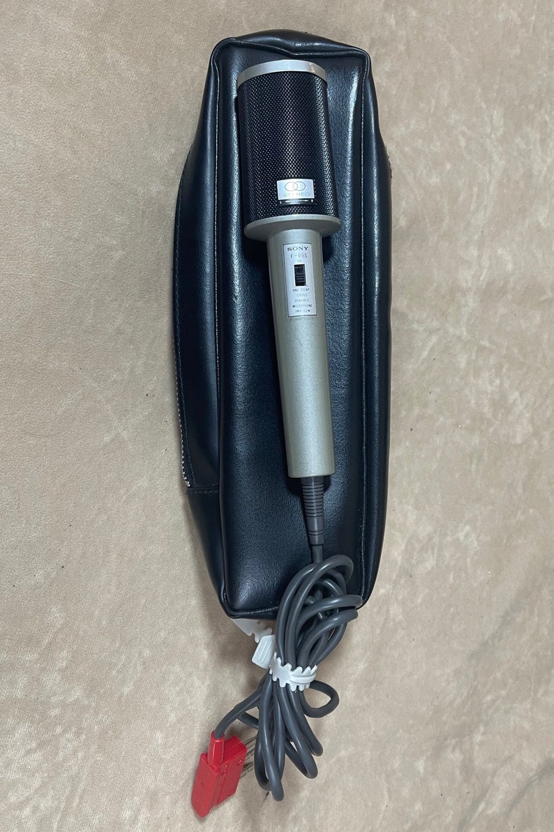 May include: A silver Sony F-915 microphone with a black mesh grille and a black leather carrying case. The microphone has a switch for on/off and a volume control knob.