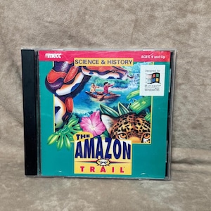 May include: A CD-ROM case for "The Amazon Trail" educational game. The cover features a vibrant illustration of a snake, jaguar, and people in a boat, with the text "SCIENCE & HISTORY" and "AGES 8 and Up". Designed for Microsoft Windows 95.