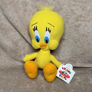 May include: A yellow plush Tweety Bird toy with blue eyes, orange beak, and black eyelashes. The plush toy has a tag that reads "Looney Tunes". The toy is sitting on a brown surface.