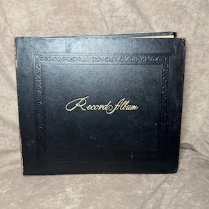 May include: A black record album with gold lettering that reads "Record Album". The album has an ornate border design and is closed. The album is sitting on a tan surface.