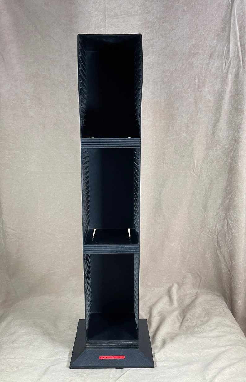 May include: A tall, black CD or DVD storage tower with multiple shelves. The tower has a rectangular base and a red label at the bottom. The tower is made of a dark material and has a vertical design.