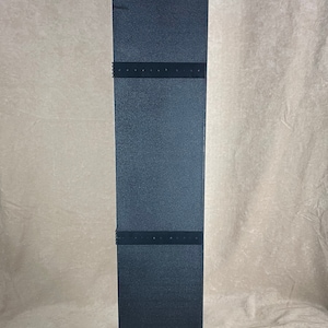 May include: A tall, black, rectangular object with a textured surface. The object has a square base and is divided into three sections by horizontal black bands. A red label is visible on the base.