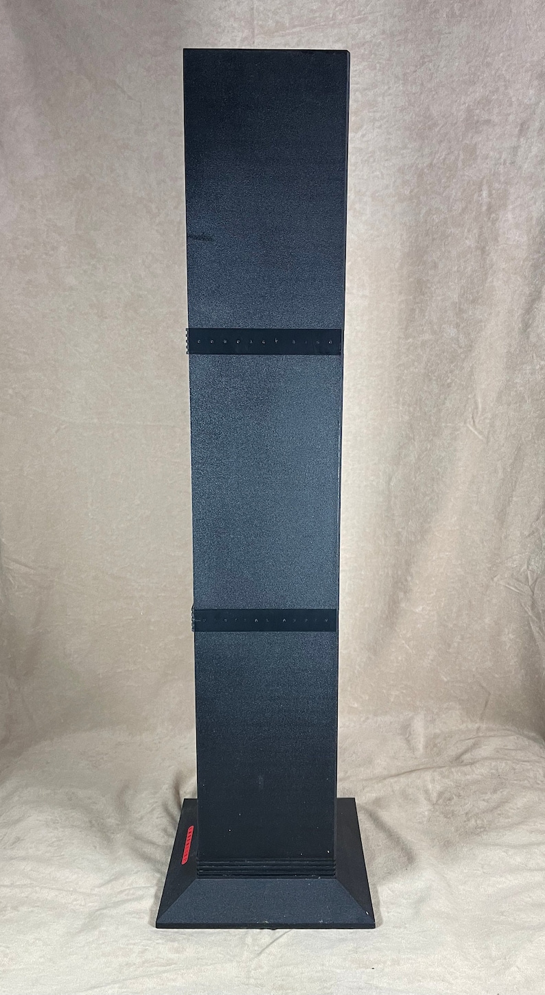 May include: A tall, black, rectangular object with a textured surface. The object has a square base and is divided into three sections by horizontal black bands. A red label is visible on the base.