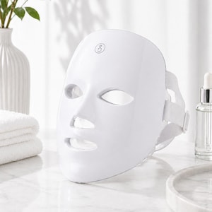 May include: White LED light therapy face mask with eye, nose, and mouth openings. The mask has a strap for secure wear and a small logo on the forehead area. A bottle and a marble tray are also visible.