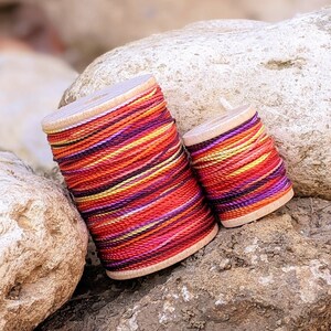 May include: Two spools of colorful embroidery floss, one larger than the other, are stacked on top of rocks. The floss is a mix of red, orange, yellow, purple, and white.