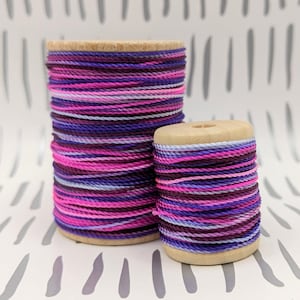 May include: Two spools of colorful embroidery floss, one larger and one smaller. The floss is a mix of pink, purple, and white.