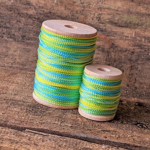 May include: Two spools of green, blue, and yellow striped thread wound around wooden bobbins. The spools are sitting on a wooden surface.