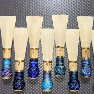 May include: A collection of nine oboe reeds, each featuring a natural wood blade and a decorative, hand-wrapped base. The bases are adorned with blue, teal, and purple threads, accented with gold-colored metal rings. The reeds are arranged against a gray background.