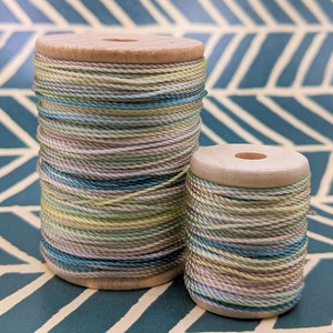 May include: Two spools of multi-colored embroidery floss, one larger than the other, with a light brown wooden base. The floss is a mix of light green, yellow, and blue.