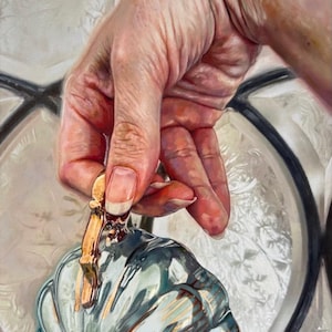 May include: A hand holds a decorative, teal-colored glass pumpkin with gold accents. The pumpkin has a white, scalloped rim and a gold stem. The background features a patterned, white surface and a black and white grid design.