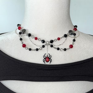 Puede incluir: Un collar de varias capas con cuentas negras y rojas, cadenas plateadas y un dije de araña colgante con una piedra preciosa roja.