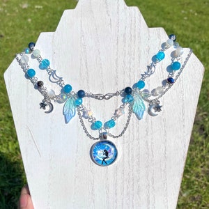 May include: A layered necklace with a silver pendant featuring a fairy sitting on a crescent moon. The necklace is made with blue and white beads, silver chains, and silver charms of stars and moons.