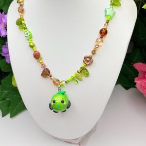 May include: A beaded necklace with a green pendant shaped like a cartoon character. The necklace features a variety of beads in shades of green, brown, and clear, with leaf and star-shaped accents. The pendant has black accents and a green stem.