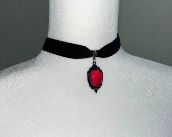 Puede incluir: Collar gargantilla de terciopelo negro con un colgante ovalado rojo. El colgante está enmarcado por un marco decorativo de metal negro. El collar se exhibe en un maniquí blanco.