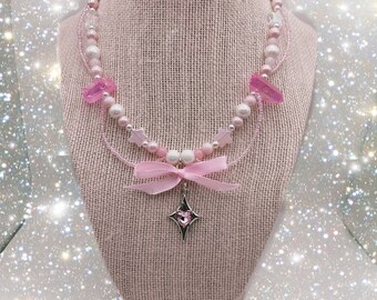 May include: A delicate pink and white layered necklace with a star charm and pink crystal accents. The necklace is displayed on a pink fabric mannequin.