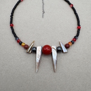 May include: A beaded necklace featuring a black beaded strand with red and pink accents. The necklace has a central design of a red sphere, two shell-like pendants, and smaller beads in various colors.