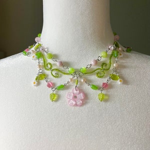 May include: A delicate multi-strand necklace with green and pink beads, silver chain, and small flower accents. The necklace features a larger pink flower charm in the centre.