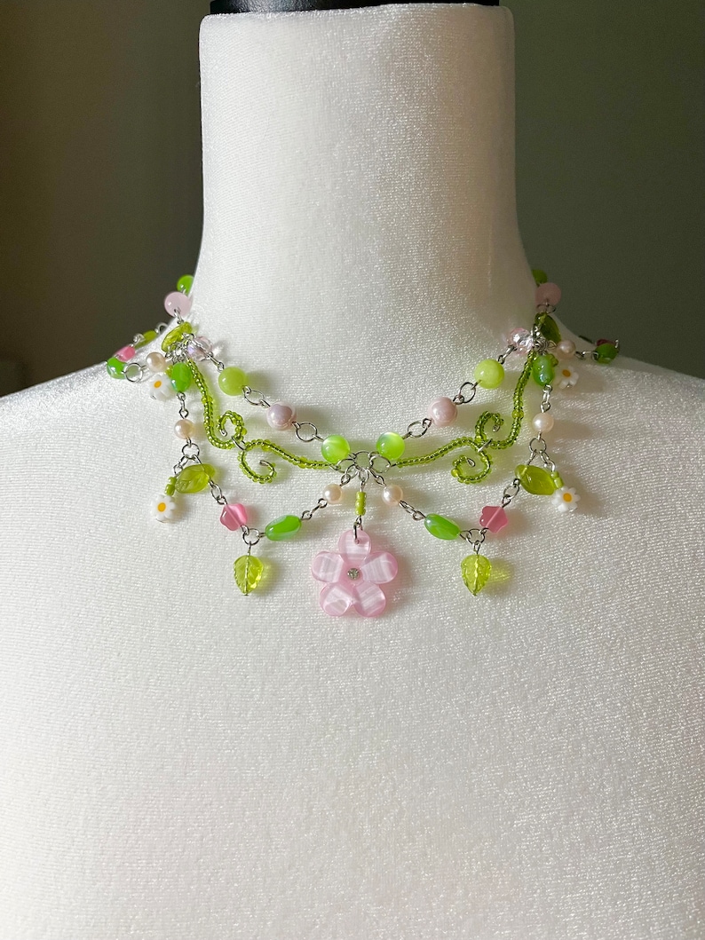 May include: A delicate multi-strand necklace with green and pink beads, silver chain, and small flower accents. The necklace features a larger pink flower charm in the centre.