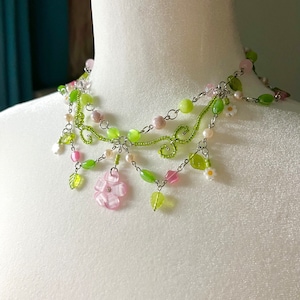 May include: A multi-strand necklace featuring green and pink beads, a silver chain, and small flower charms. The necklace has a delicate floral design with leaves and flowers.