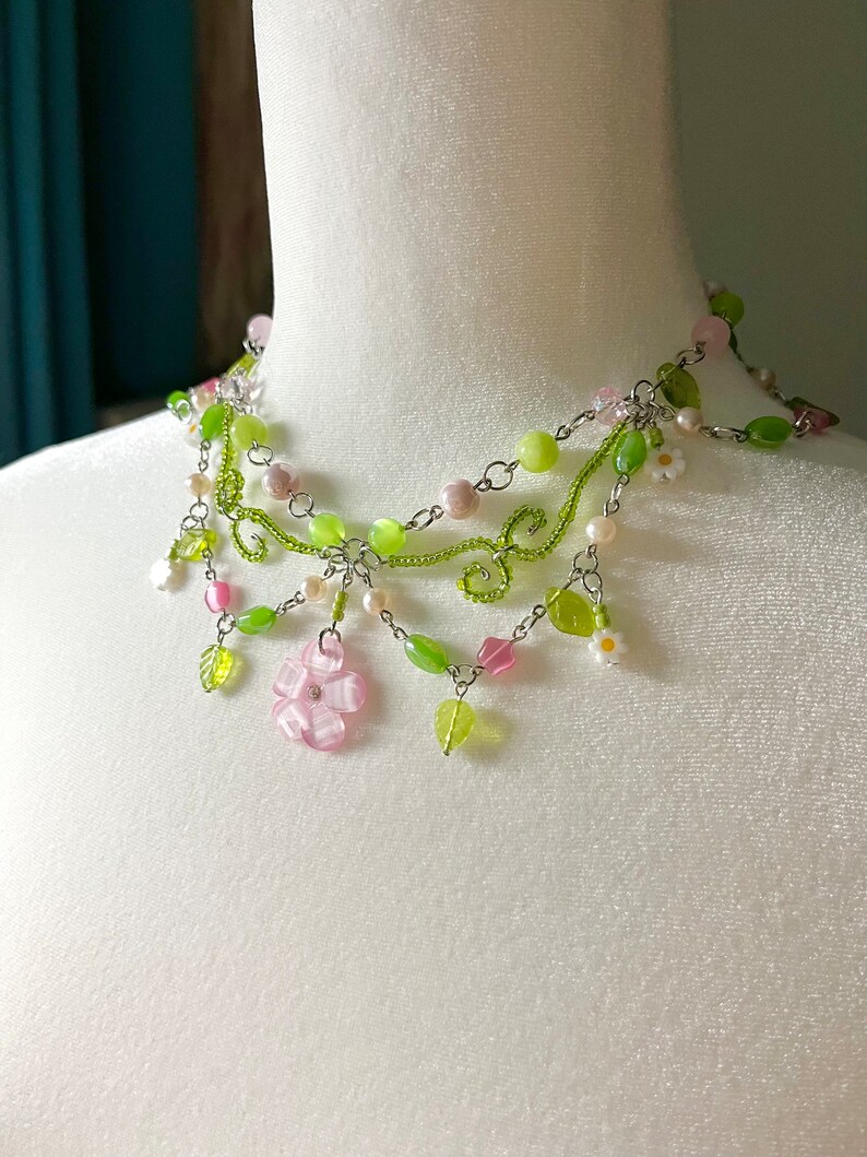 May include: A multi-strand necklace featuring green and pink beads, a silver chain, and small flower charms. The necklace has a delicate floral design with leaves and flowers.