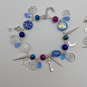K&ouml;nnte beinhalten: Ein silbernes Armband mit blauen, violetten und wei&szlig;en Perlen und silbernen Anh&auml;ngern, darunter Schl&uuml;ssel, Sterne, Monde und Spiralen.